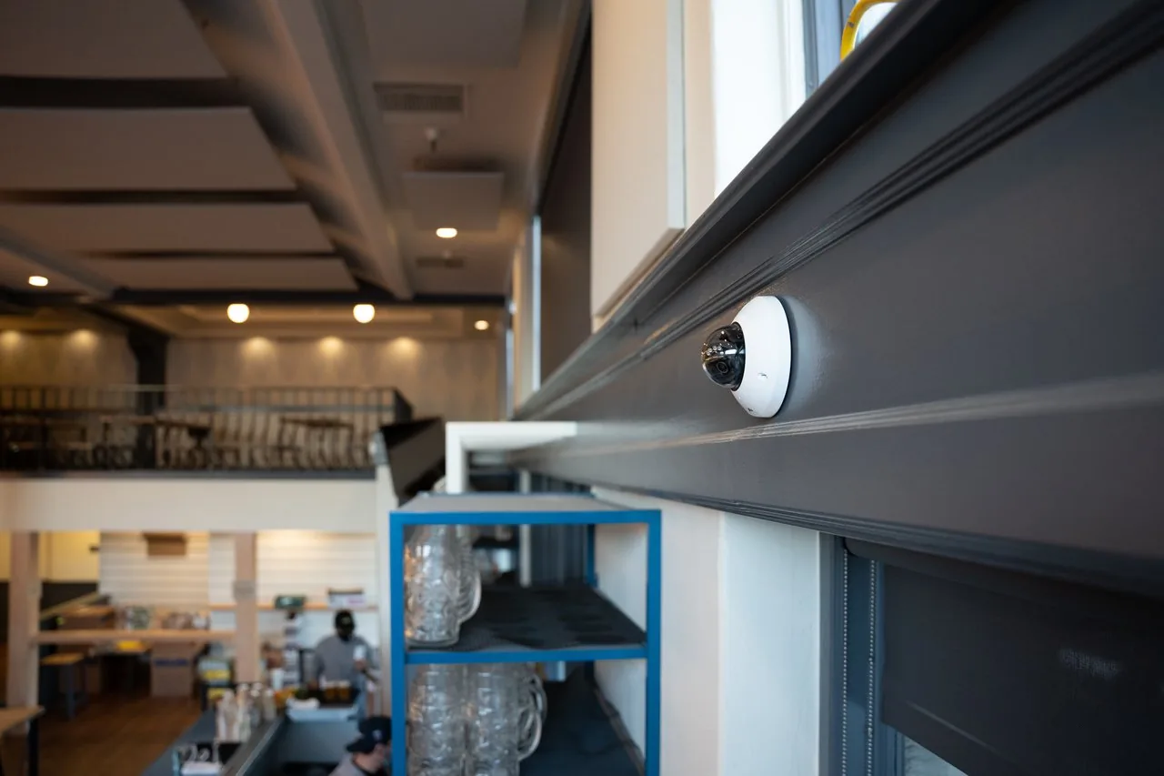 Wall-mounted dome security camera on a gray beam overlooking a multi-level restaurant interior with a mezzanine.