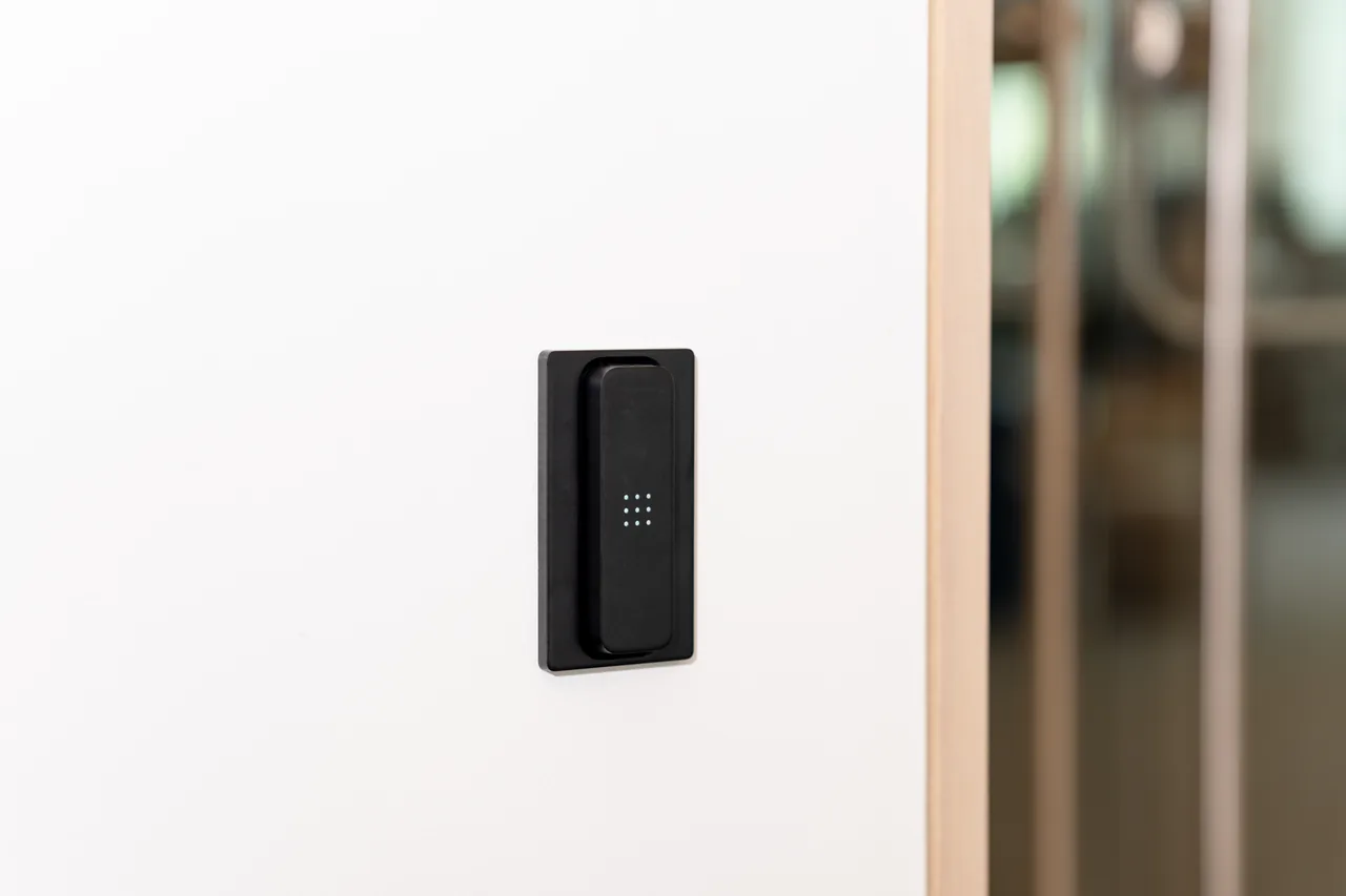 Black wall-mounted reader with dot-matrix sensor, part of a door access control system for office entry beside a glass.