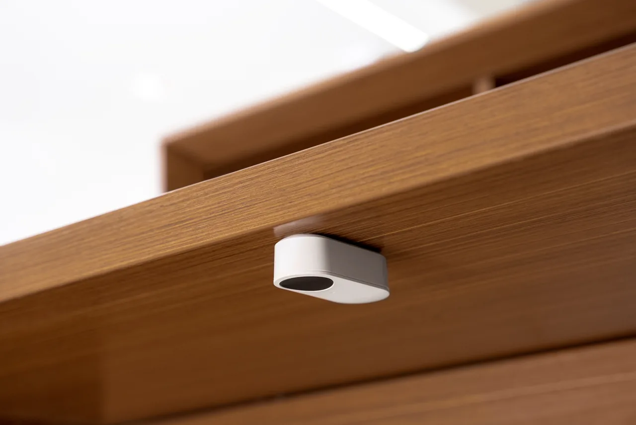 Small white panic button mounted discreetly beneath a warm wood-grain desk surface, facing outward for easy activation.