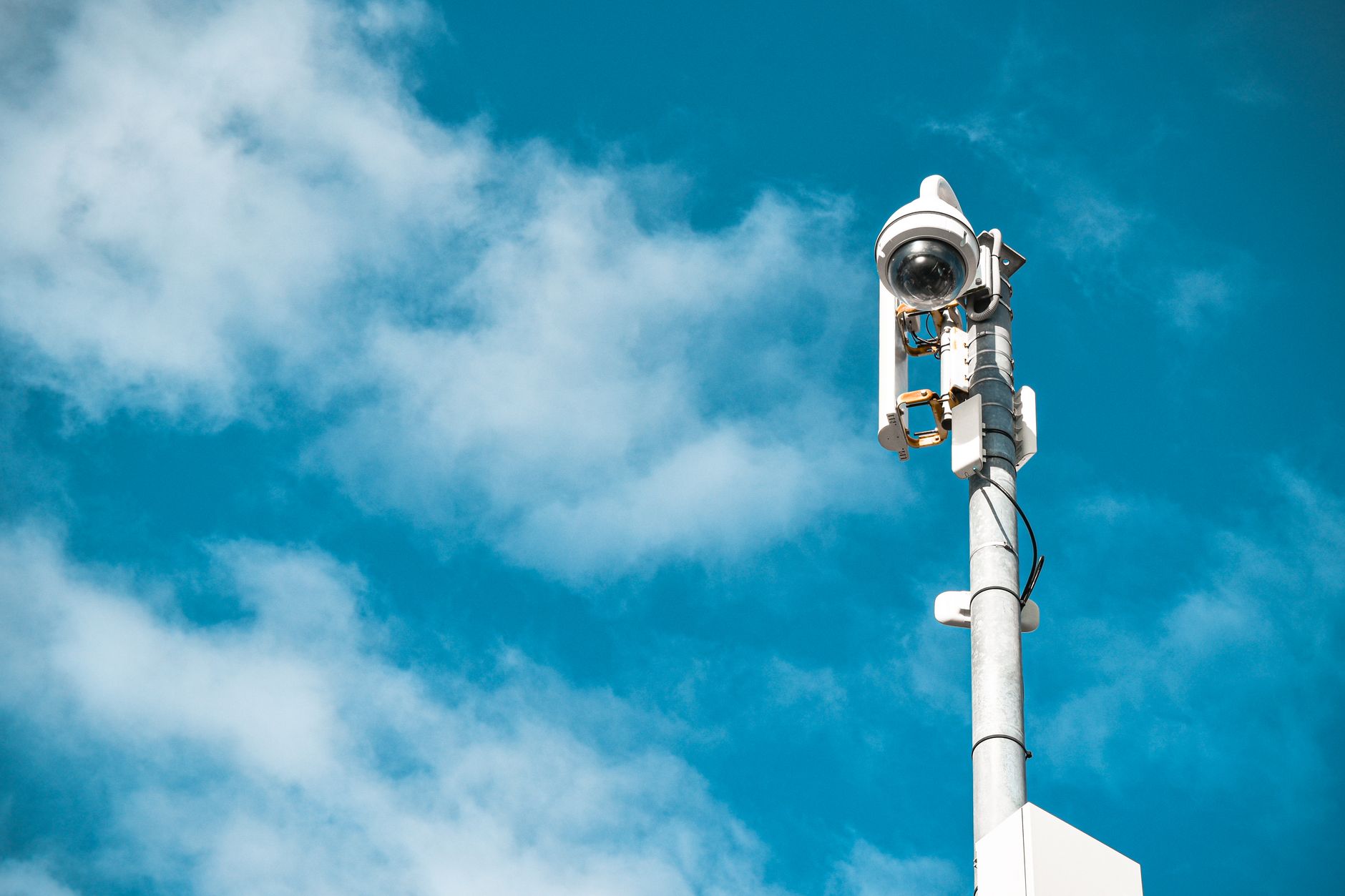 Outdoor dome security camera on a tall pole running CCTV software against a bright blue cloudy sky.