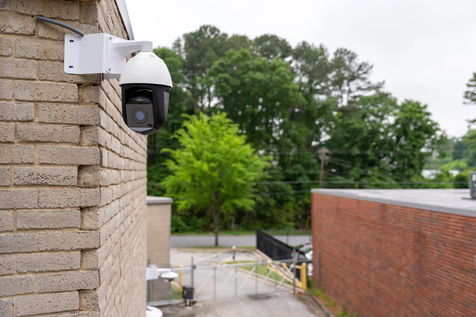 PTZ security camera mounted on a building exterior covering a fenced warehouse yard and vehicle gate
